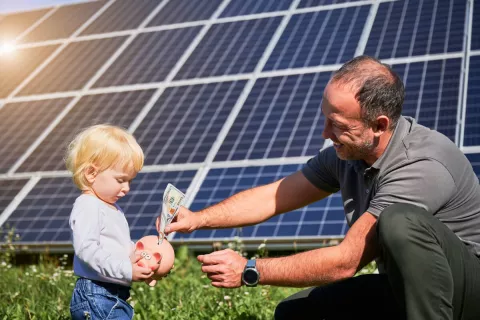 Opa steckt Geld ins Sparschwein des Enkelkindes und im Hintergrund ist eine große Solaranlage zu sehen.